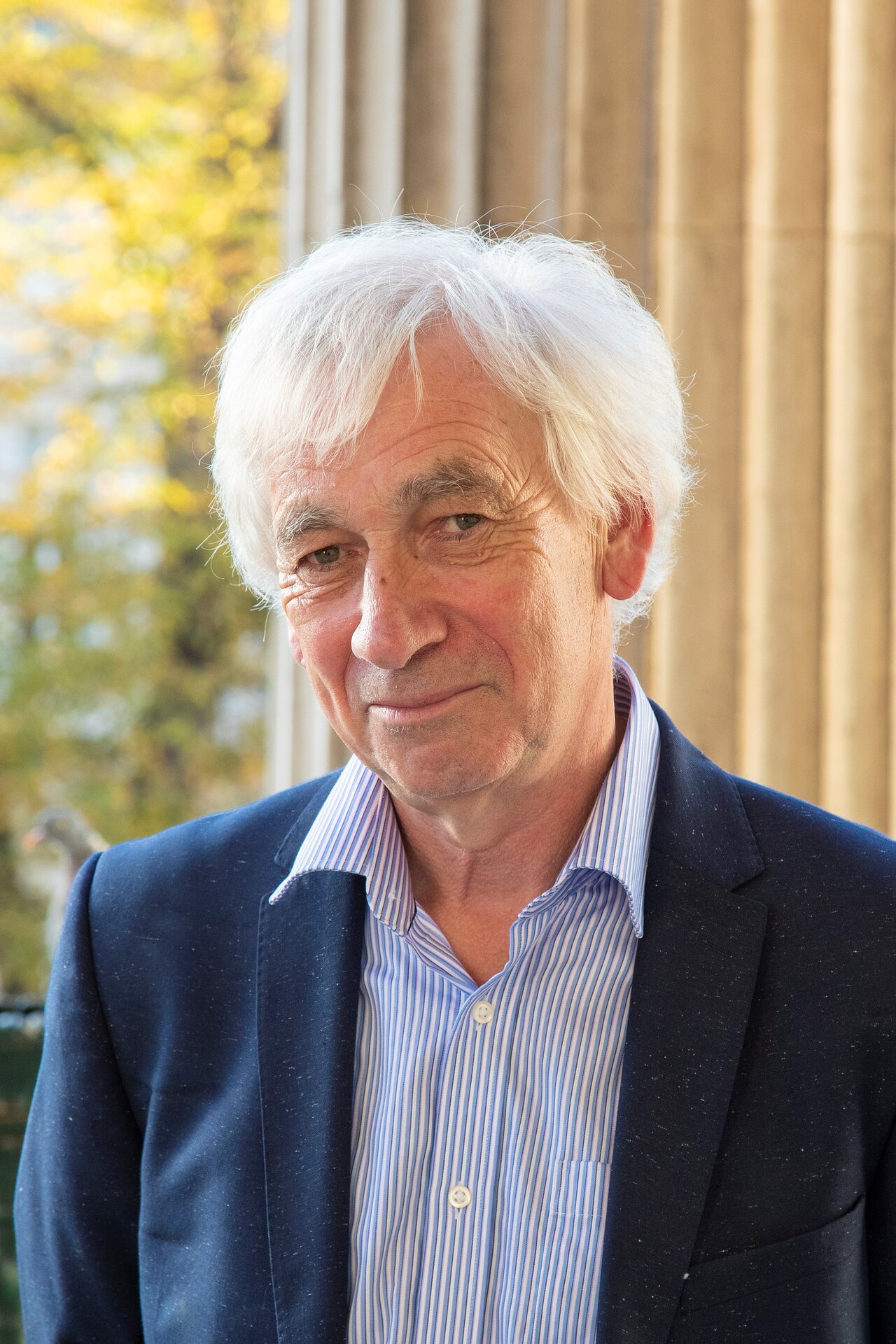 Older man with white hair wearing dark blazer and striped shirt, photographed indoors with curtained window and autumn foliage visible behind.