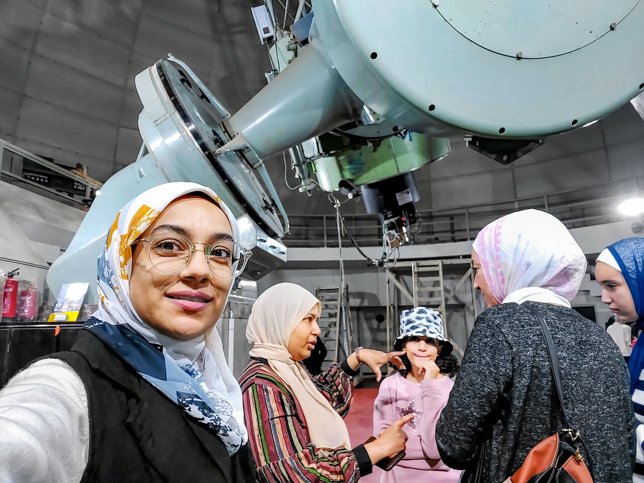 Woman wearing glasses and headscarf smiles at camera inside observatory dome with telescope equipment and group of visitors behind her.