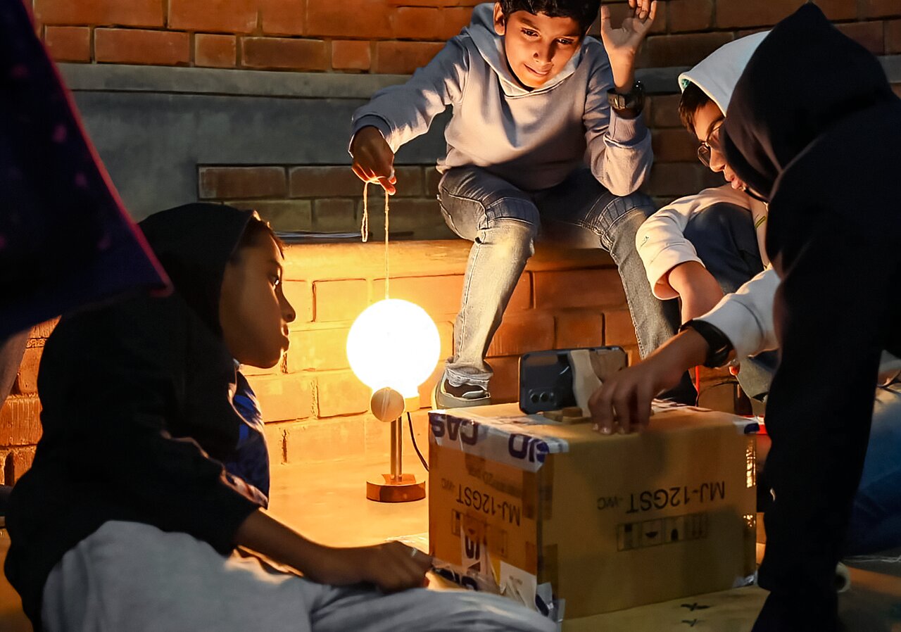 Children gather around a glowing spherical model suspended on a stand, exploring a hands-on astronomy demonstration indoors.