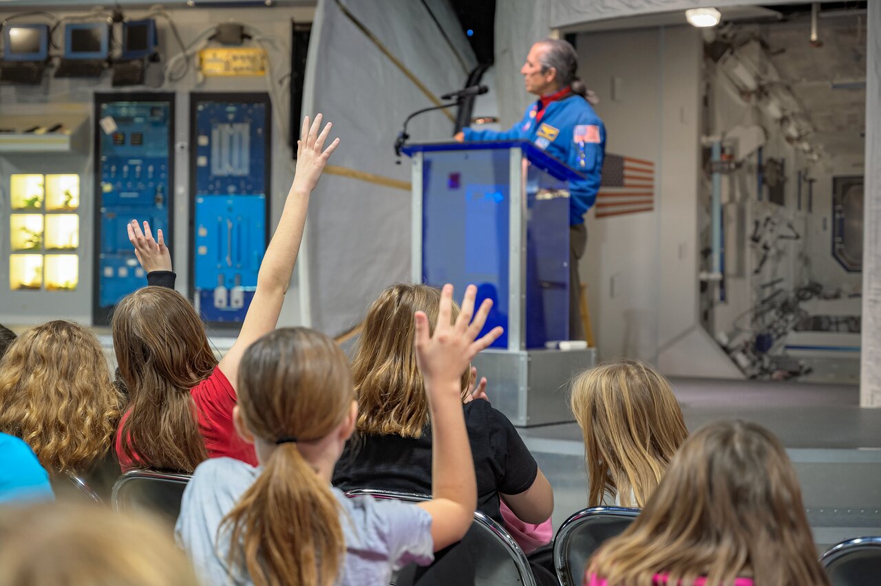 Children raise hands eagerly while listening to a speaker in a blue flight suit at a podium inside a spacecraft simulator facility.