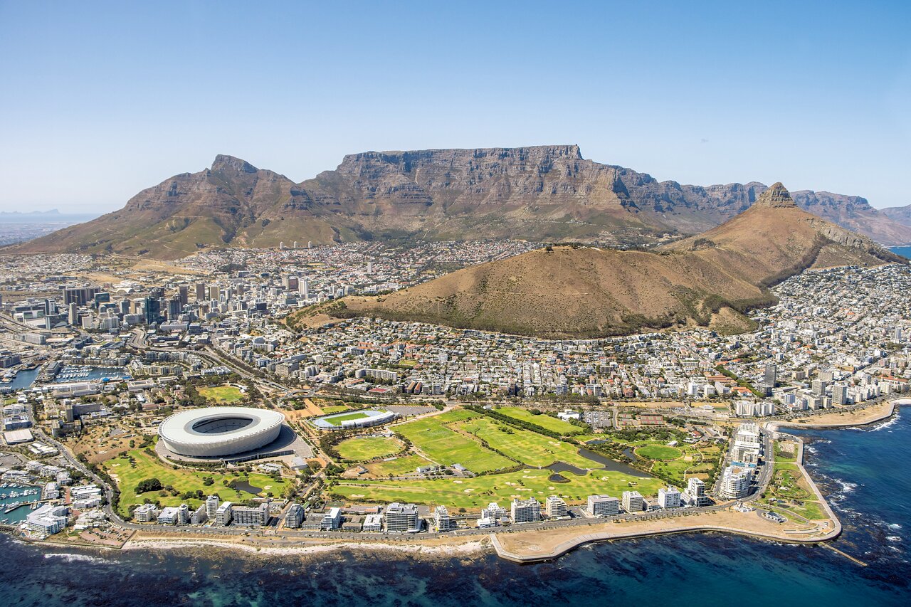 Aerial view of a coastal city with distinctive flat-topped mountain, surrounding peaks, modern stadium, and turquoise ocean waters under clear sky.