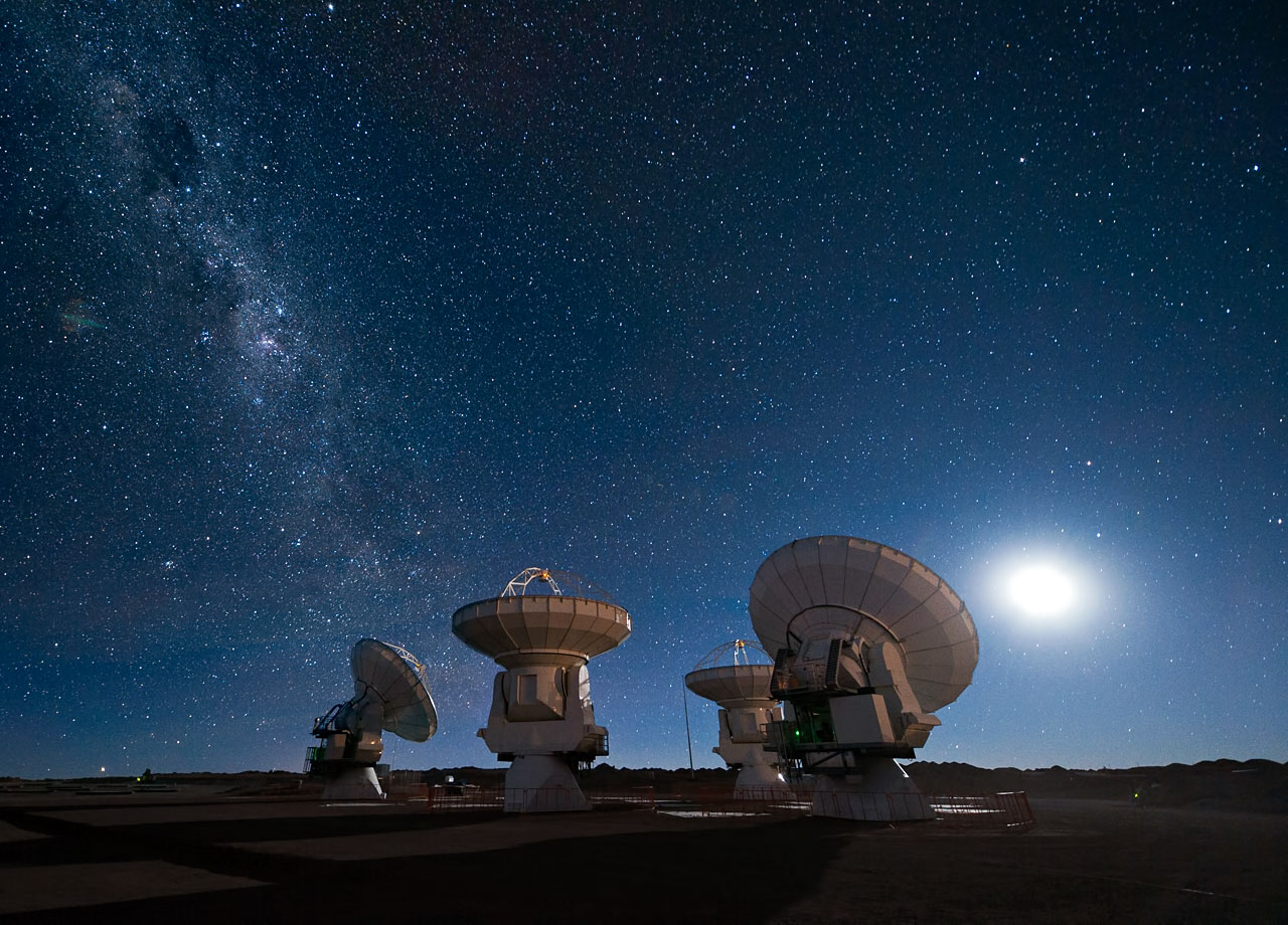 Four antennas of the Atacama Large Millimeter/submillimeter Array (ALMA) gaze up at the star-filled night sky. The Moon lights the scene on the right, while the band of the Milky Way stretches across the upper left.